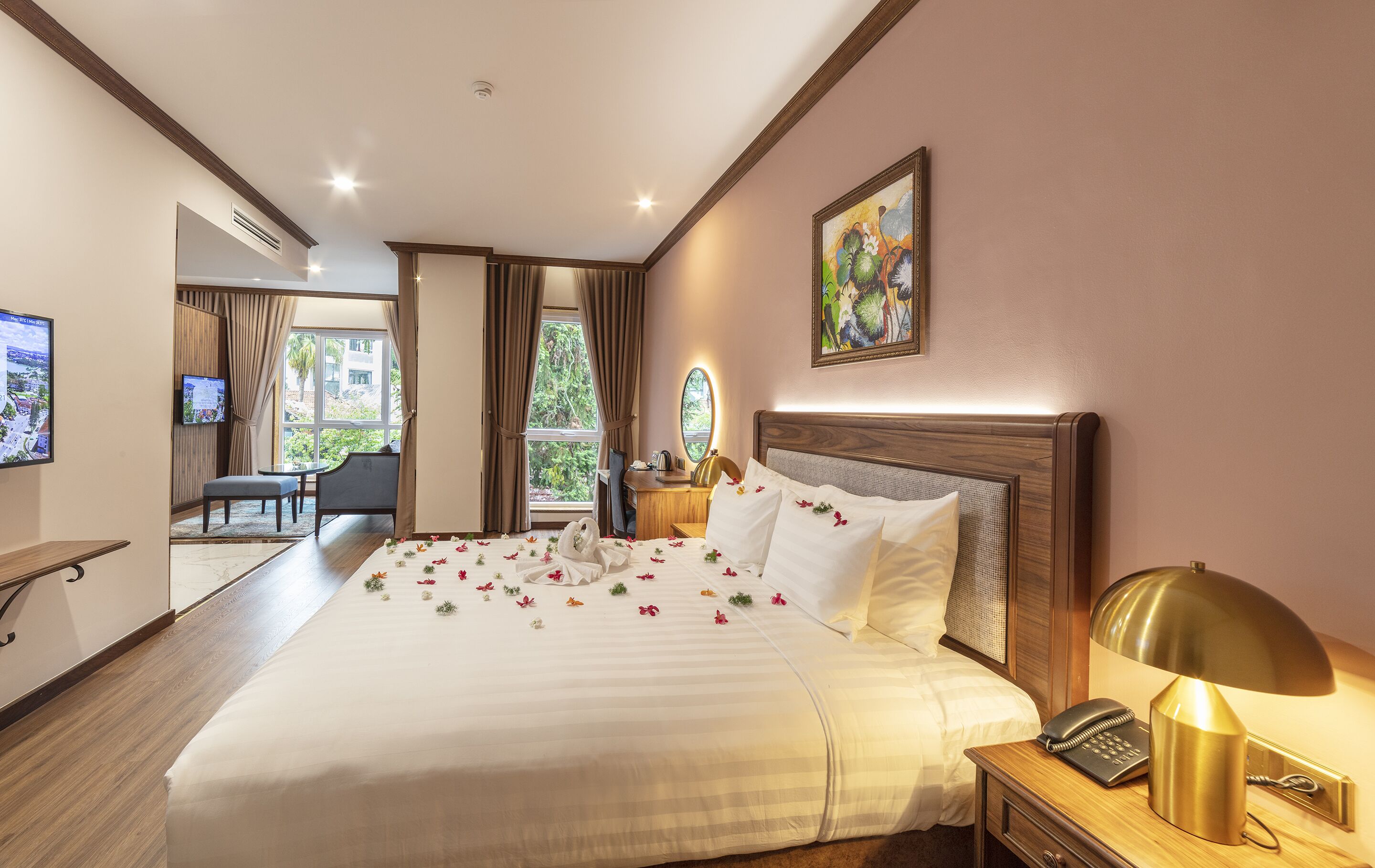 A hotel room with a large bed, a bedside table with a lamp, a TV, a desk with a chair, and a view of greenery outside.