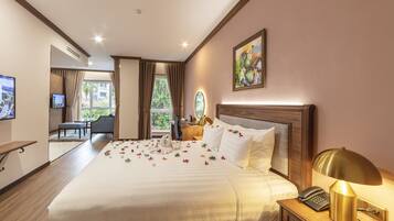 A hotel room with a large bed, a bedside table with a lamp, a TV, a desk with a chair, and a view of greenery outside.