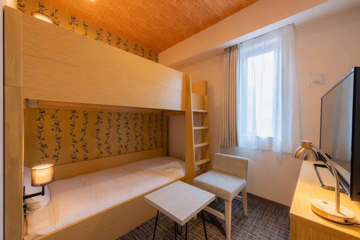 Habitación de hotel compacta con literas, un escritorio pequeño con lámpara, una silla y un televisor.