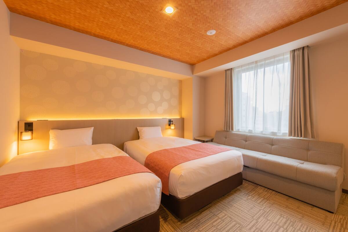 Habitación de hotel con dos camas, un sofá, un papel tapiz estampado y una ventana con cortinas.