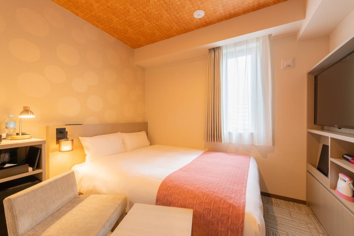 Una habitación de hotel con cama, escritorio con lámpara, televisión y ventana con cortinas.