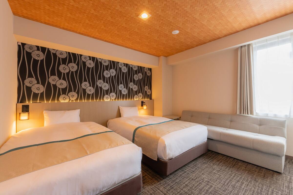 Habitación de hotel con dos camas, un sofá, un papel tapiz estampado y una lámpara en el techo.