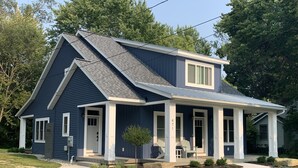 A two-story house with a gabled roof, a covered porch with chairs, and a small tree in the front yard. - Beautiful newer construction 2021 home in town! (Saugatuck)
