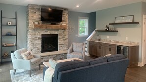 A modern living room with a stone fireplace, a flat-screen TV, and a kitchen area with a marble countertop. - Beautiful newer construction 2021 home in town! (Saugatuck)