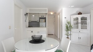 A modern kitchen with white cabinetry, a glass dining table, and a potted plant. - Cala Blanca, low, 2 bedrooms, by Bookindenia (Dénia)