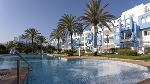 A swimming pool with a clear blue water surface, surrounded by palm trees and a building with blue and white architecture. - Cala Blanca, low, 2 bedrooms, by Bookindenia (Dénia)