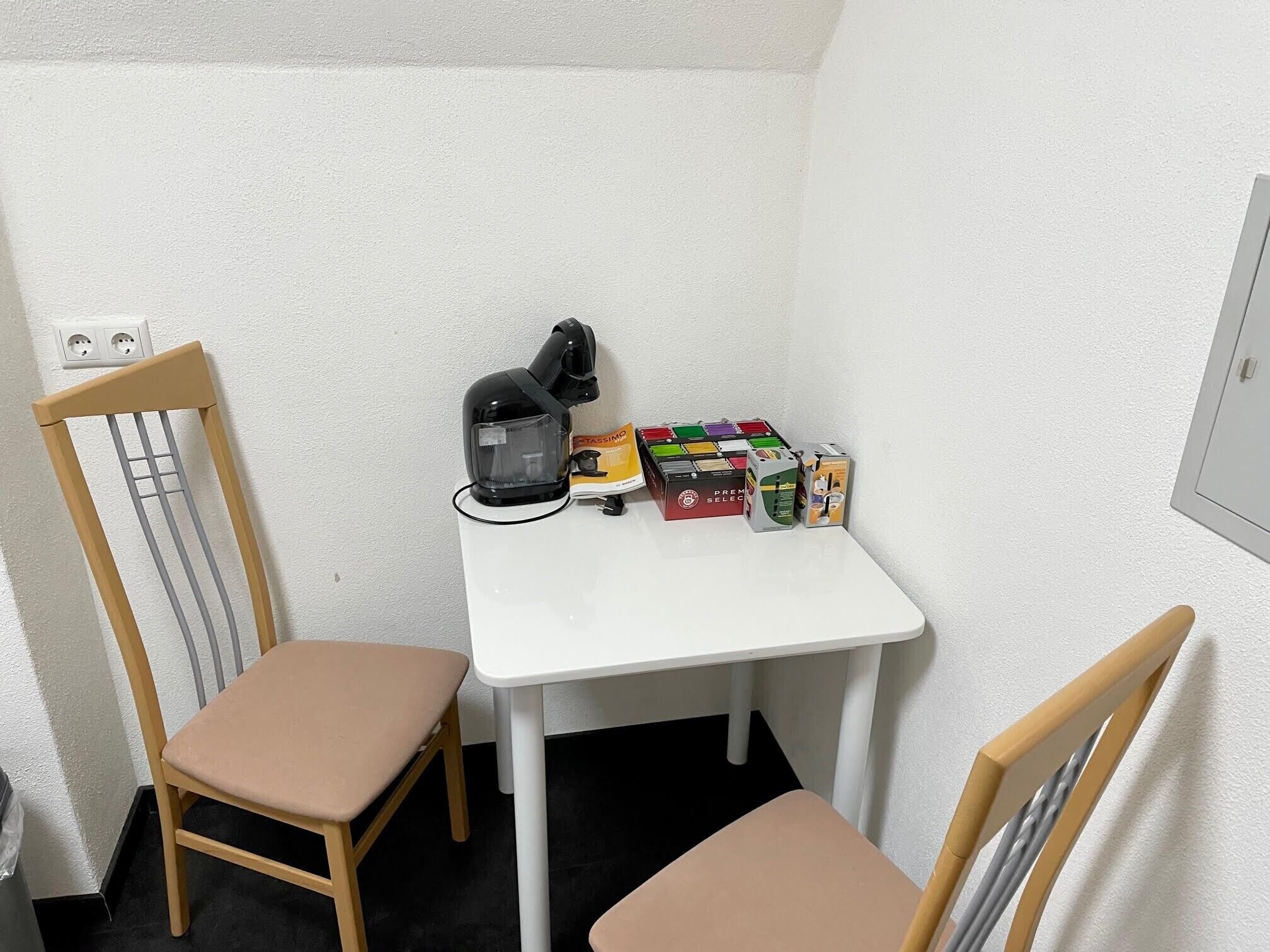 A small room with a white table, two chairs, a black kettle, and a box of TASSIMO pens.