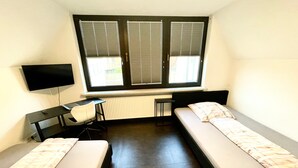 Ein Zimmer mit zwei Einzelbetten, einem Schreibtisch, einem Stuhl, einem Fernseher und einem Fenster mit Jalousien.