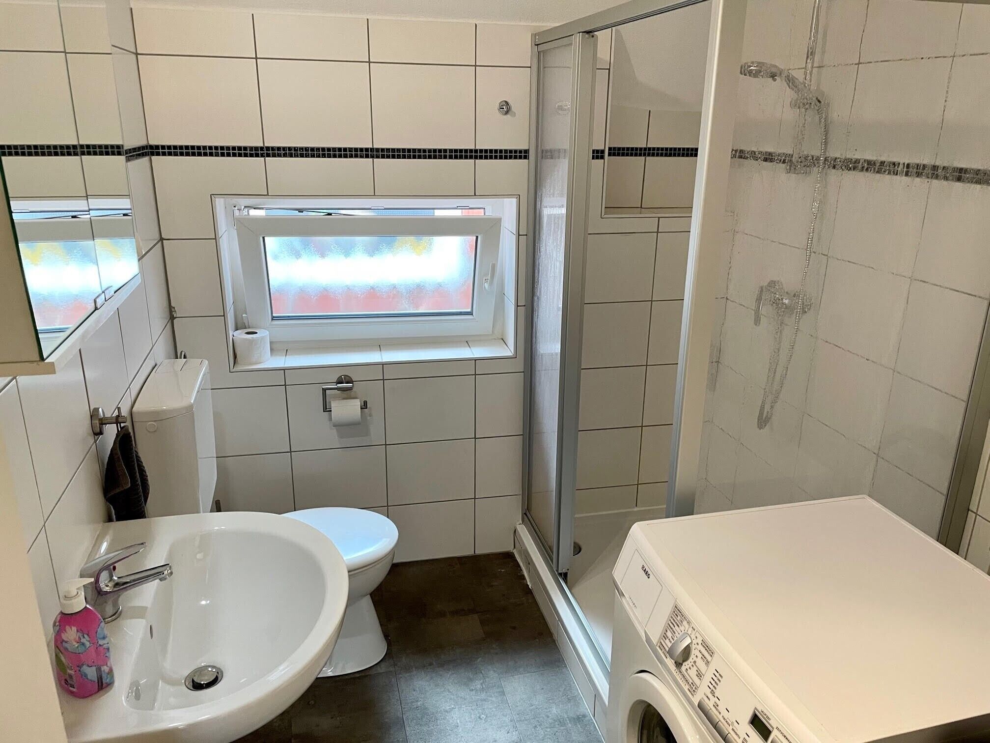 A bathroom with a toilet, sink, shower, and a window.