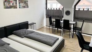 Une chambre moderne équipée d’un lit, d’un bureau et d’une chaise, d’une horloge au mur et d’une fenêtre donnant sur des immeubles.