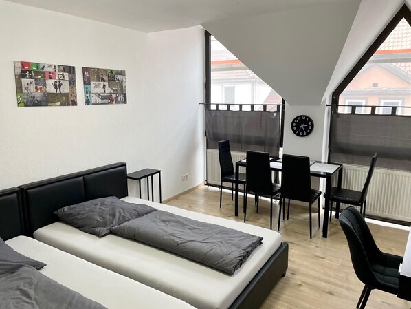 A modern room with a bed, a desk, and a chair, a clock on the wall, and a window with a view of buildings.