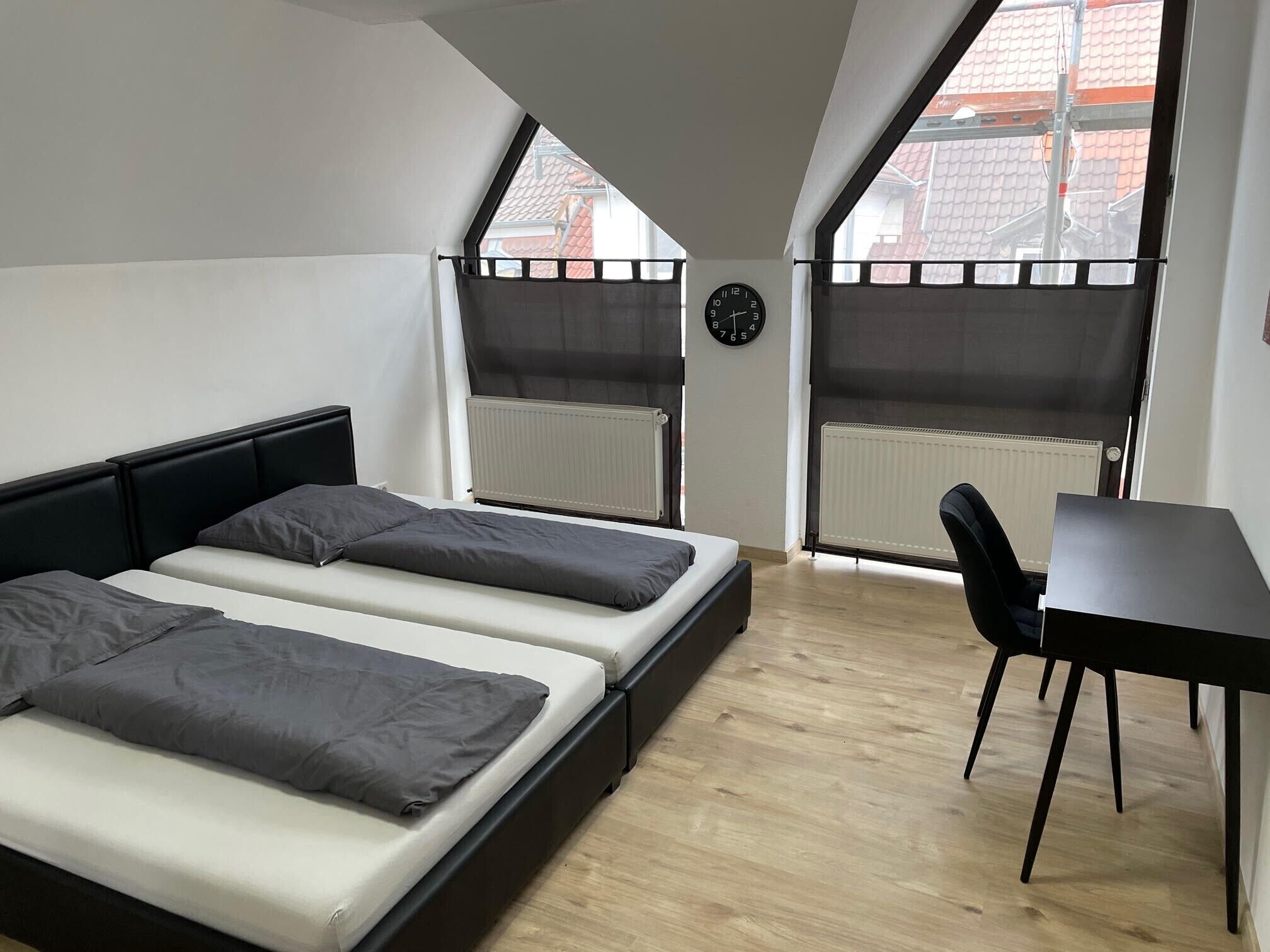 Ein Zimmer mit einem Bett, einem Schreibtisch und einem Stuhl. Zwei Fenster mit Jalousien, ein Heizkörper und eine Uhr an der Wand sind zu sehen.