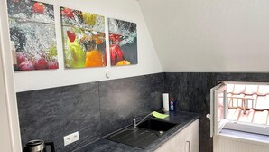 Une cuisine moderne avec des plans de travail sombres, un évier et une bouilloire. Trois tableaux colorés représentant des fruits sont accrochés au mur.