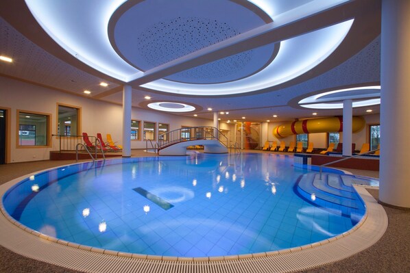 Una amplia piscina interior con una escalera central, varias zonas de descanso y un diseño de techo moderno.