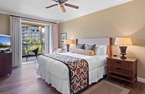 A hotel room with a bed, two bedside tables with lamps, a ceiling fan, a TV, and a balcony with a view of a building and palm trees.