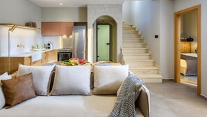 A modern living room with a sofa, a coffee table with fruits, a kitchen area with cabinets, and a staircase. - Caldera Theros Villas (Hersonissos)