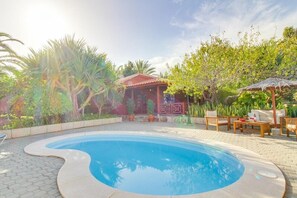 Una piscina con borde curvo, rodeada por un patio empedrado. Hay una terraza de madera con mobiliario exterior y una zona de estar cubierta.