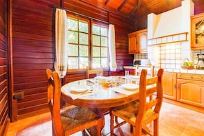 Interior de una cabaña de madera con una mesa de comedor para cuatro, sillas de madera, una zona de cocina con armarios y un amplio ventanal con cortinas.