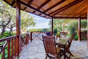 Un área de comedor al aire libre techada con suelo de piedra, mobiliario de madera y vistas a una piscina y vegetación.