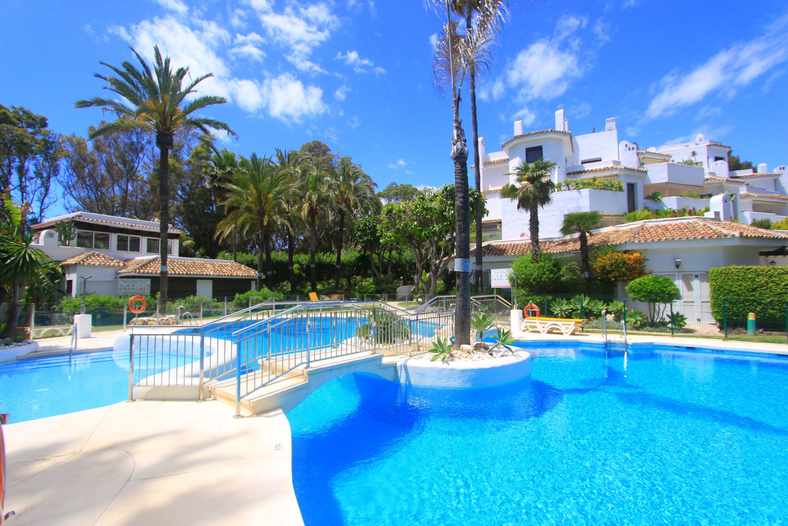 A swimming pool with a surrounding deck, palm trees, and a building with white walls and red-tiled roofs in the background.