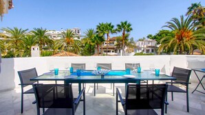 An outdoor dining area with a glass table and chairs, surrounded by palm trees and buildings in the background.