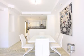 Un espace de repas moderne avec une table et des chaises blanches, une cuisine en arrière-plan et un tableau représentant un tigre accroché au mur.