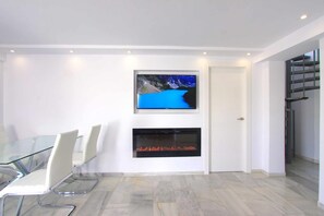 A modern interior with a glass table, white chairs, a mounted TV, and a fireplace. - 1094 Luxurious Golden Beach Duplex Penthouse (Marbella)