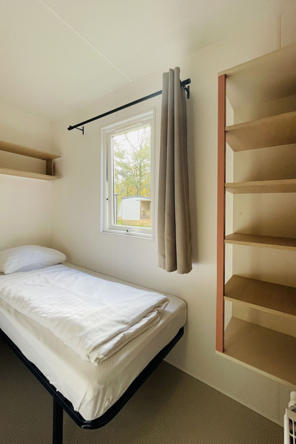 A single bed with a headboard, a window with curtains, and a built-in shelving unit.