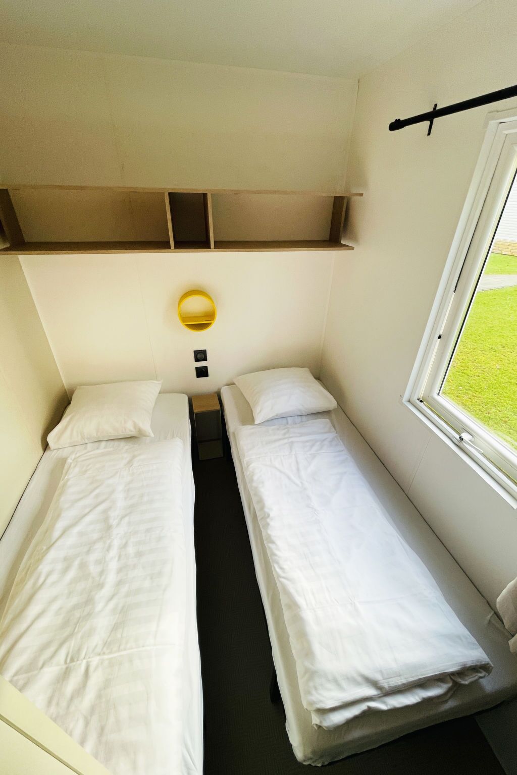 A small, compact room with two single beds, a small shelf above the beds, a bedside table, a wall-mounted light, and a window with a view of a grassy area.