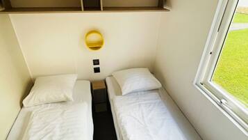 A small, compact room with two single beds, a small shelf above the beds, a bedside table, a wall-mounted light, and a window with a view of a grassy area.