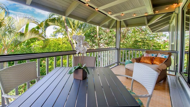 A covered outdoor area with a table and chairs, surrounded by lush greenery and a view of the sky.