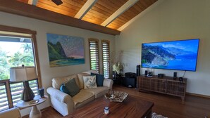 A living room with a wooden ceiling, a beige sofa, a coffee table, a TV on the wall, and a painting of a landscape.