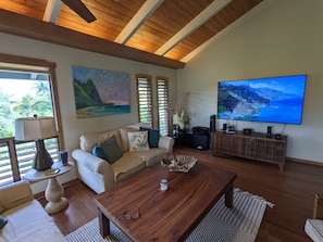 A living room with a wooden ceiling, a beige sofa, a coffee table, a TV on the wall, and a painting of a landscape. - Executive Family 4BR Princeville Home by beaches & the park w/AC, SPA and Bikes (Princeville)