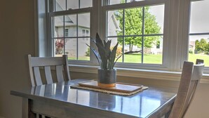 A dining table with a potted plant, two chairs, and a view of a residential area through large windows.