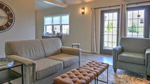 A spacious living room with a grey sofa, a matching armchair, and a brown ottoman. A dining area is visible in the background with a table and chairs.