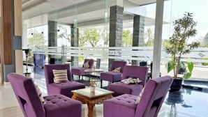 A modern lobby with purple armchairs arranged around a glass coffee table, large windows offering a view of the outdoors, and a stone pillar. - Harmoni Garut (Garut)