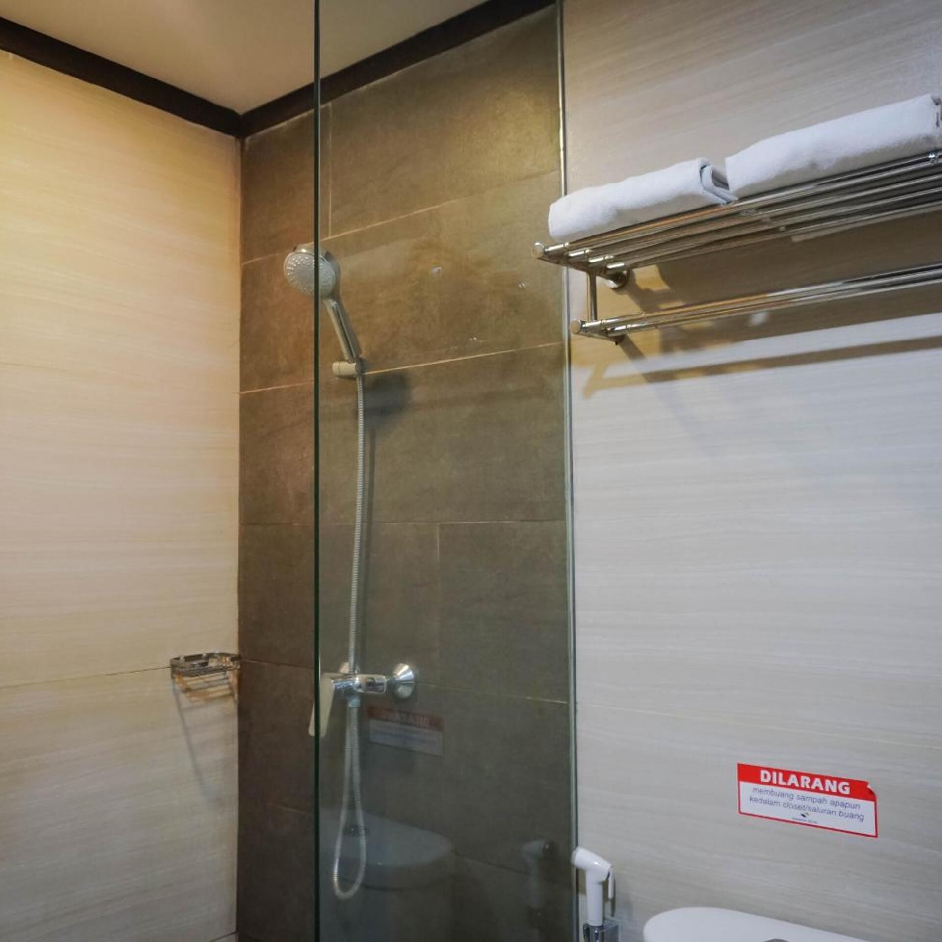 A glass-enclosed shower with a handheld showerhead, a towel rack, and a toilet with a 'DILARANG' sign.