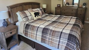 Een slaapkamer met een geruit sprei, een houten hoofdbord, een nachtkastje met lamp, een dressoir en een wanddecoratie van een boslandschap.