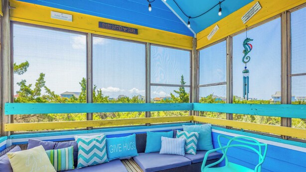 A sunroom with a blue and yellow exterior, a wicker sofa with cushions, and a teal chair. - Soundside Home w/ Private Pool, Game Room, Basketball, and Nearby Beach Access (Avon)