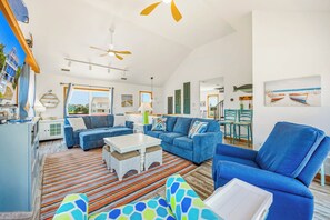 A spacious living room with a large sofa, a smaller sofa, a coffee table, and a chair. There are two ceiling fans, a window with a view, and a dining area with chairs and a table.