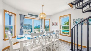 A dining area with a table set for four, white chairs, a chandelier, and large windows with a view. - Soundside Home w/ Private Pool, Game Room, Basketball, and Nearby Beach Access (Avon)
