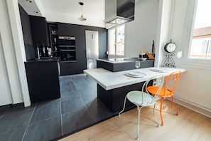 A modern kitchen with dark cabinetry, a central island, and a dining area with a table and chairs. - Appt Lumineux F3 Quartier Calme (Vichy)