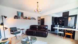 Un moderno salón con un sofá de cuero, una mesa de centro de vidrio y un escritorio con una lámpara. Hay una zona de cocina con una mesa de comedor y sillas.