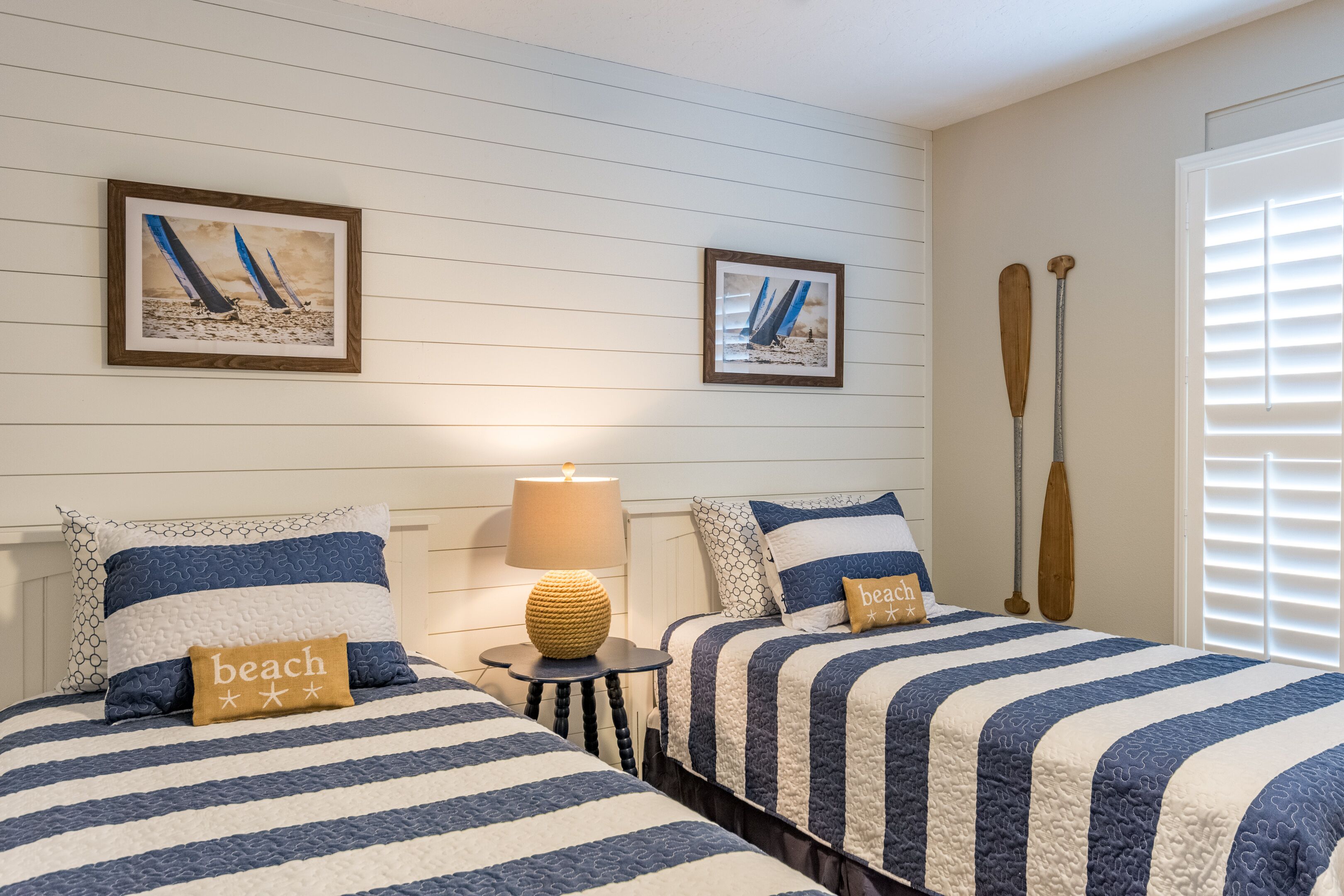 A bedroom with two beds, striped bedding, a small table with a lamp, and two framed pictures on the wall.