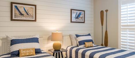 A bedroom with two beds, striped bedding, a small table with a lamp, and two framed pictures on the wall.