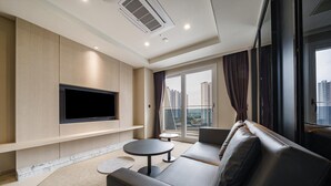 Una sala de estar moderna con un televisor de pantalla plana, un sofá, una mesita redonda y vistas a un paisaje urbano a través de amplias ventanas.