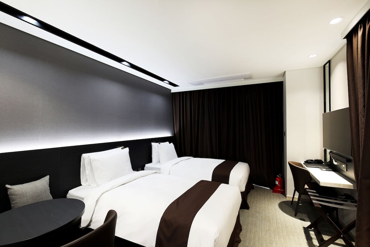A hotel room with two beds, a desk, a chair, a round table, and a fire extinguisher.
