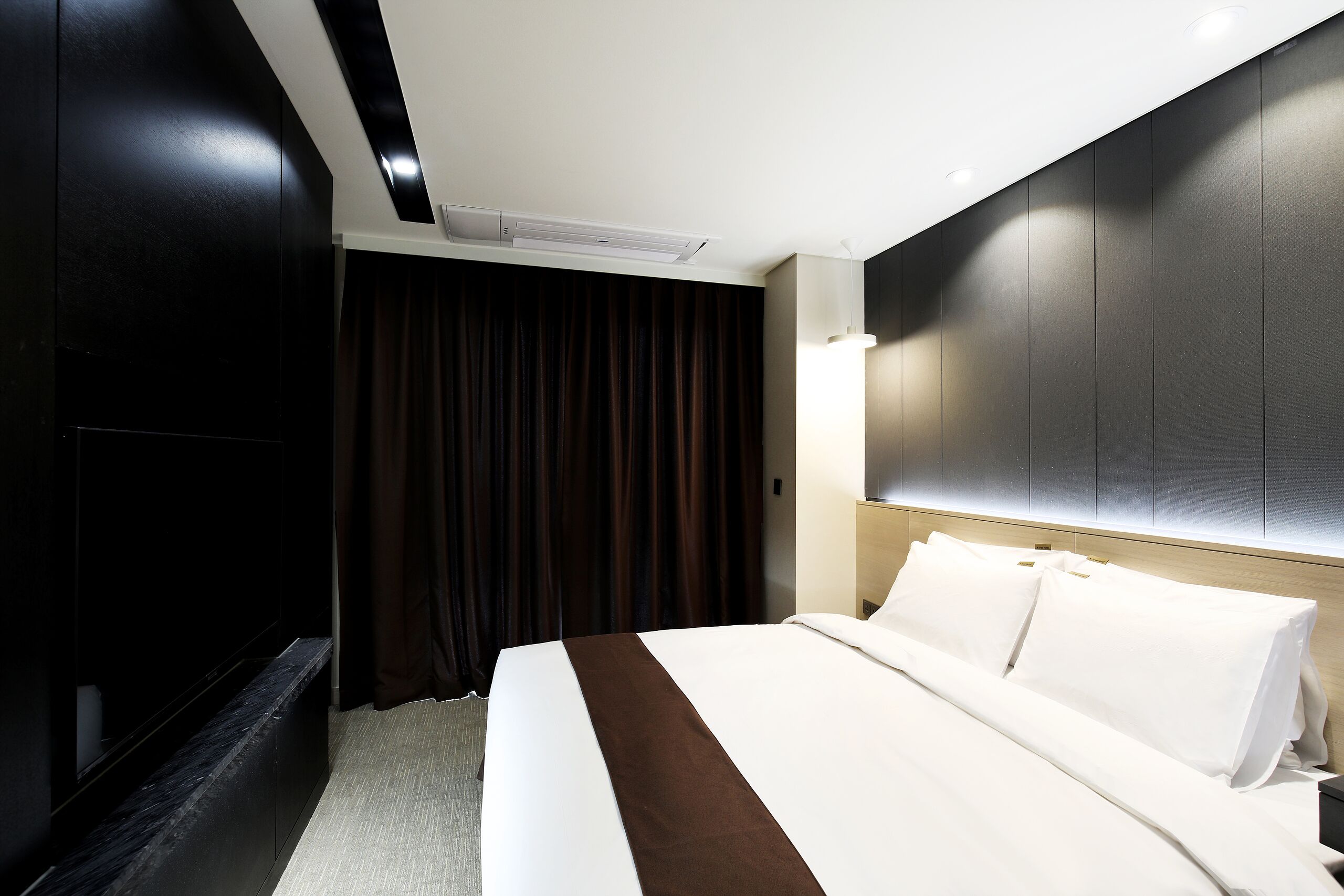 Habitación de hotel con una cama grande, cortinas oscuras, aire acondicionado instalado en el techo y un cabecero moderno.