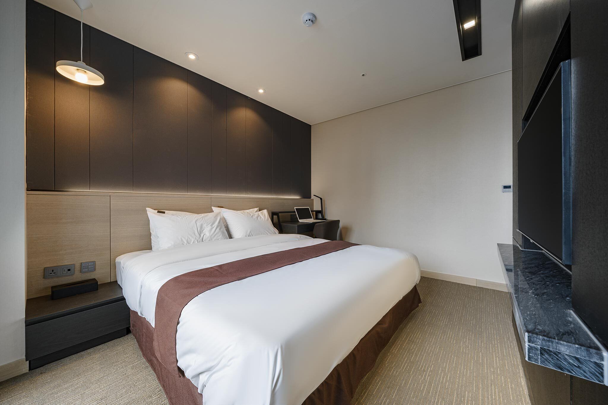 A modern hotel room with a large bed, a flat-screen TV, a desk with a laptop, and a wall-mounted TV.