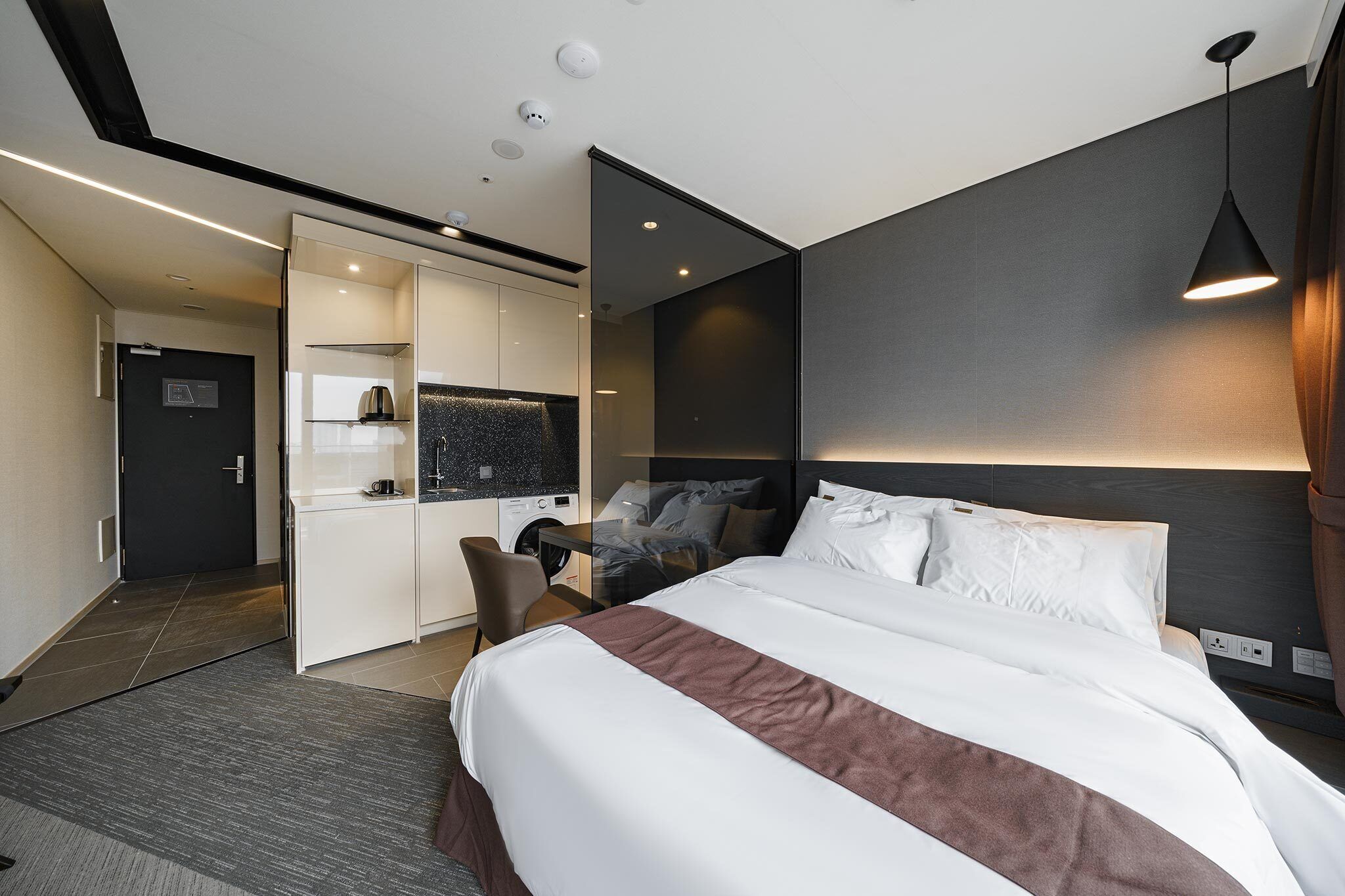 Una habitación de hotel moderna con una cama grande, un escritorio con silla, un armario empotrado y una pequeña kitchenette.